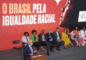 Imagem colorida mostra Lula discursando no dia 21 de março de 2023 durante assinatura de decretos que institui ações nos 20 anos das políticas de igualdade racial no Brasil. Ao fundo está escrito a mensagem: O Brasil pela igualdade racial